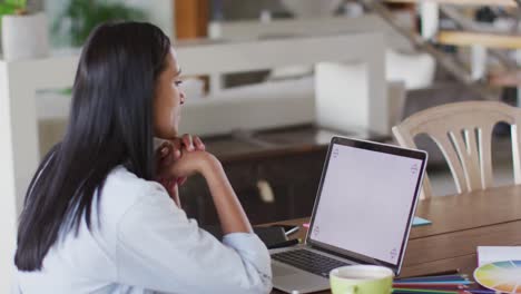 Mixed-race-woman-using-laptop-writing-in-notebook-drinking-coffee-working-from-home