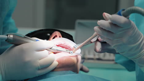 the hands of a dentist and an assistant with surgical instruments, a dental drill treat caries to a female patient. patient wearing goggles with open mouth