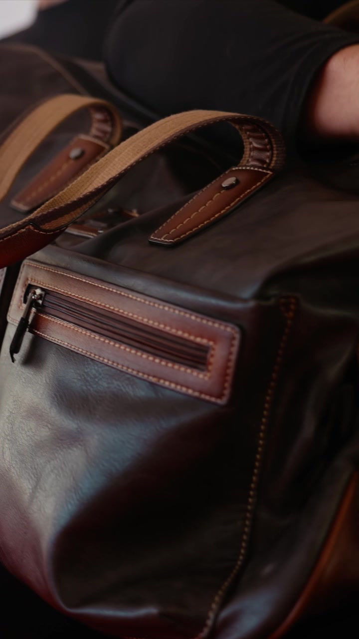 Primo piano di una persona che maneggia borse di pelle marrone scuro. La persona interagisce da vicino con la borsa, regolando le cinghie o preparandosi a spostarla all'interno. L'illuminazione evidenzia i dettagli.