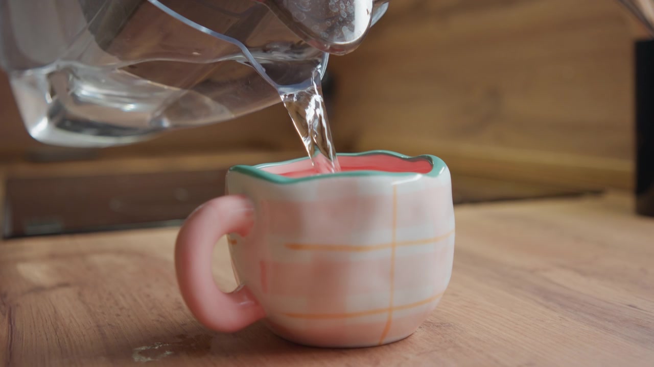 Una scena di cucina di profilo mostra una tazza smaltata rosa su un piano di lavoro in legno mentre un liquido trasparente si versa da una brocca di vetro inclinata, creando un flusso visibile, un leggero spruzzo, increspature, riflessi e un'atmosfera domestica calda.