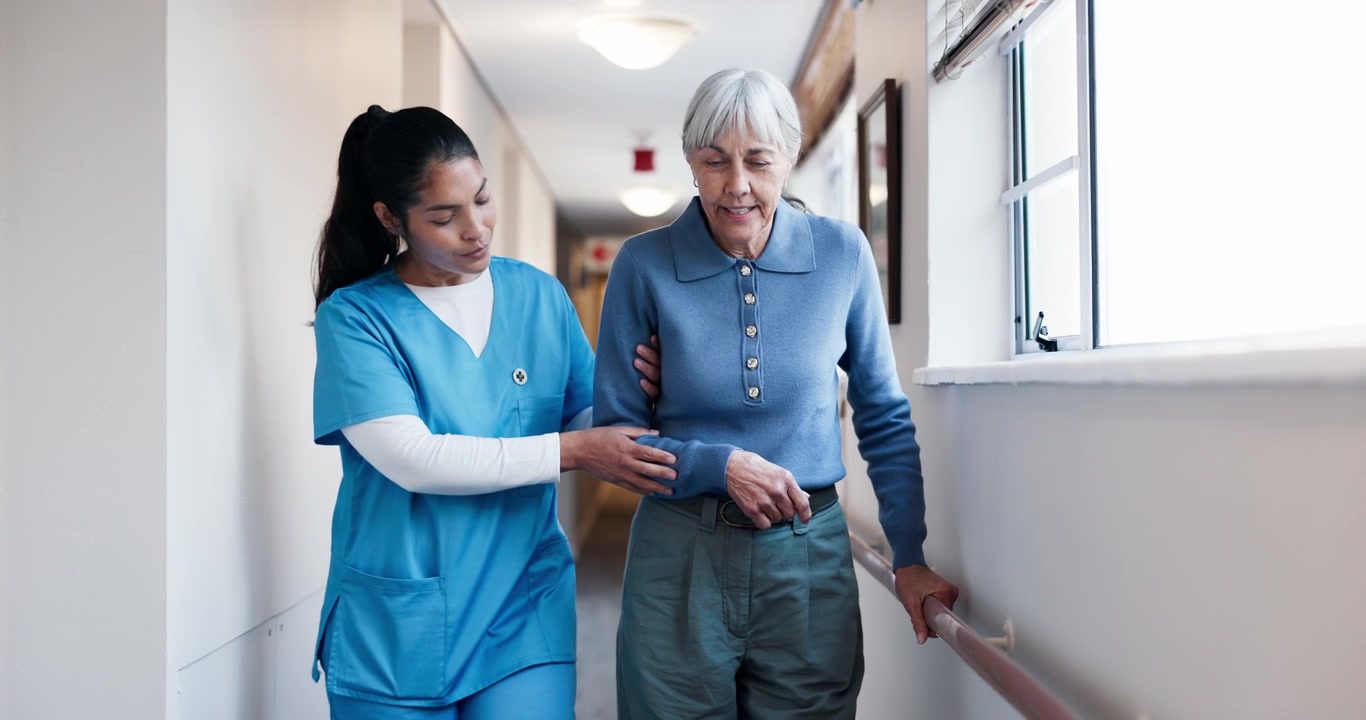 Caregiver Assisting Elderly Woman Walking Down Hospital Hallway