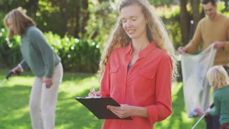 video portrait of caucasian woman with clipboard and volunteers collecting plastic waste outdoors