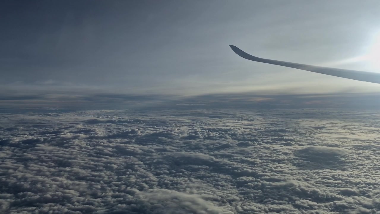 Utsikt fra vindusplass av flyvinge som flyr høyt over en tilsynelatende endeløs flate av teksturerte hvite skyer i atmosfæren