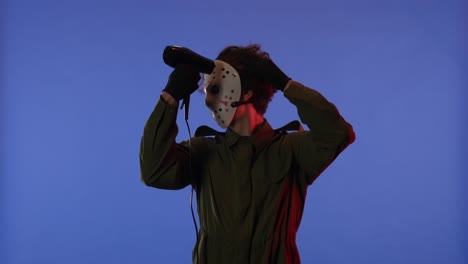 man in white mask drying hair with hair dryer on blue background with red neon light image of maniac - man in white mask drying hair with hair dryer on blue background with red neon light. image of maniac jason voorhees from friday 13. halloween celebration concept