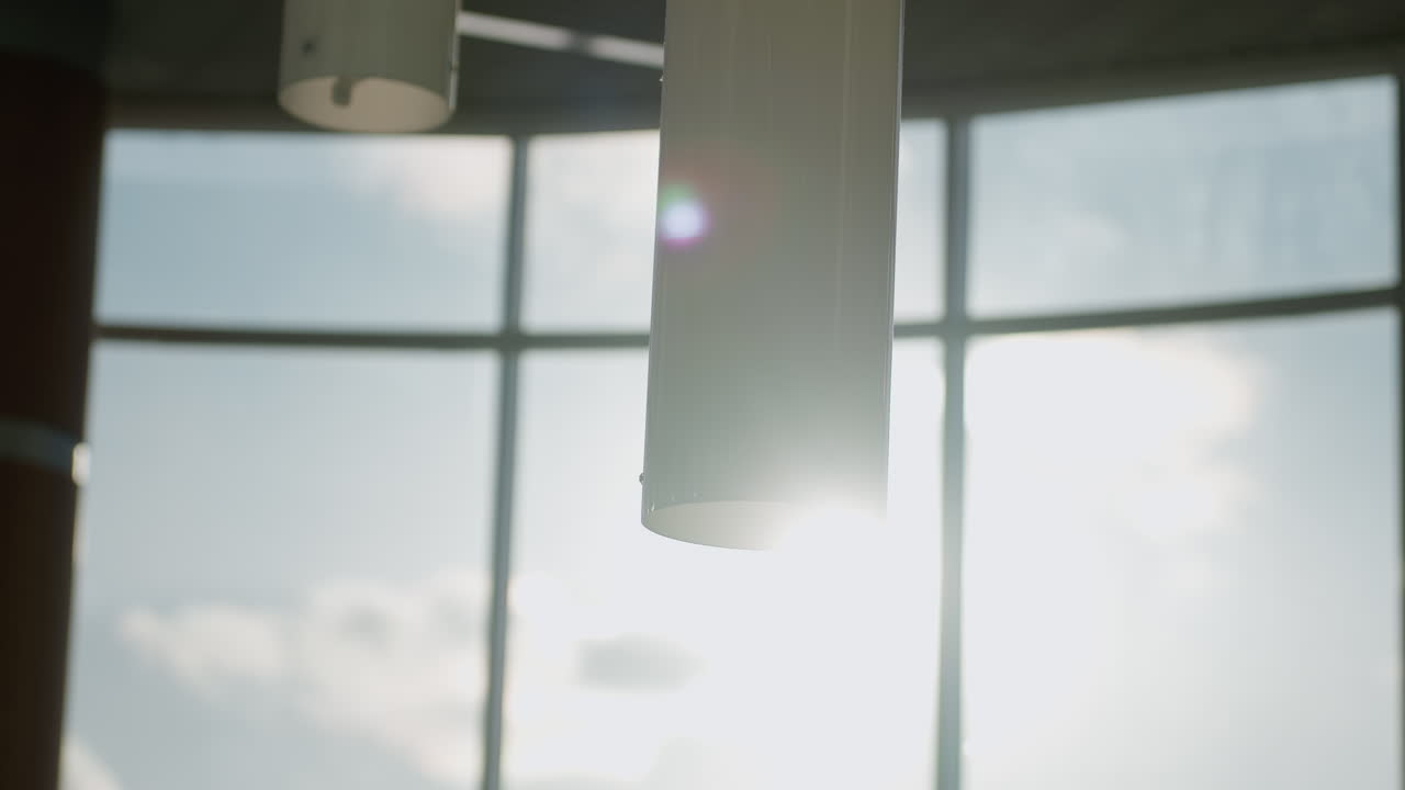Sunlight streaming through large windows casts warm glow on cylindrical hanging pendant lights in modern gallery space creating soft lens flare reflections on glass railing and wooden columns