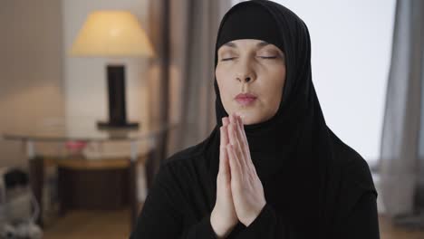 portrait of young beautiful muslim woman sitting with closed eyes and holding hands together. adult lady in black hijab praying at home. lifestyle, religion. camera moving around from right to left.