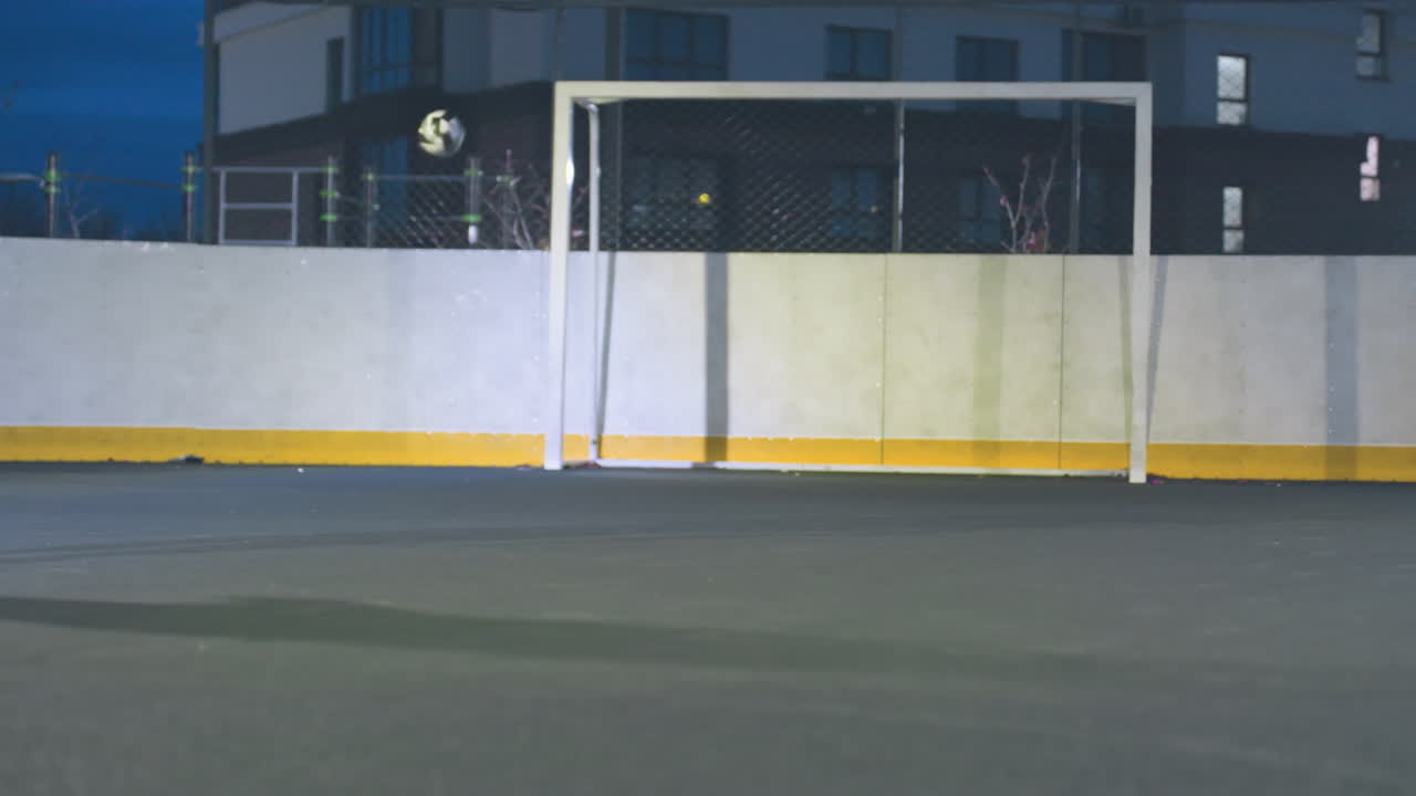 Partial view of athlete kicking soccer ball toward goal post during outdoor training, the ball hits the post and bounces out under bright stadium lights with an urban setting in the background