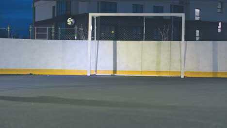 partial view of athlete kicking soccer ball toward goal post during outdoor training, the ball hits the post and bounces out under bright stadium lights with an urban setting in the background