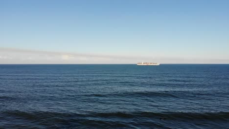 a large cargo ship sails through large, open waters off the coast to a nearby beach town under a baby blue sky that harbours low hanging fluffy clouds