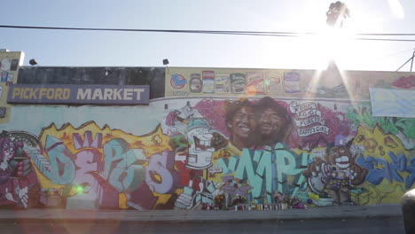 low angle wide shot of kobe gigi bryant memorial mural, pickford market in la – car drives by in slow motion, sun flare and palm tree in background