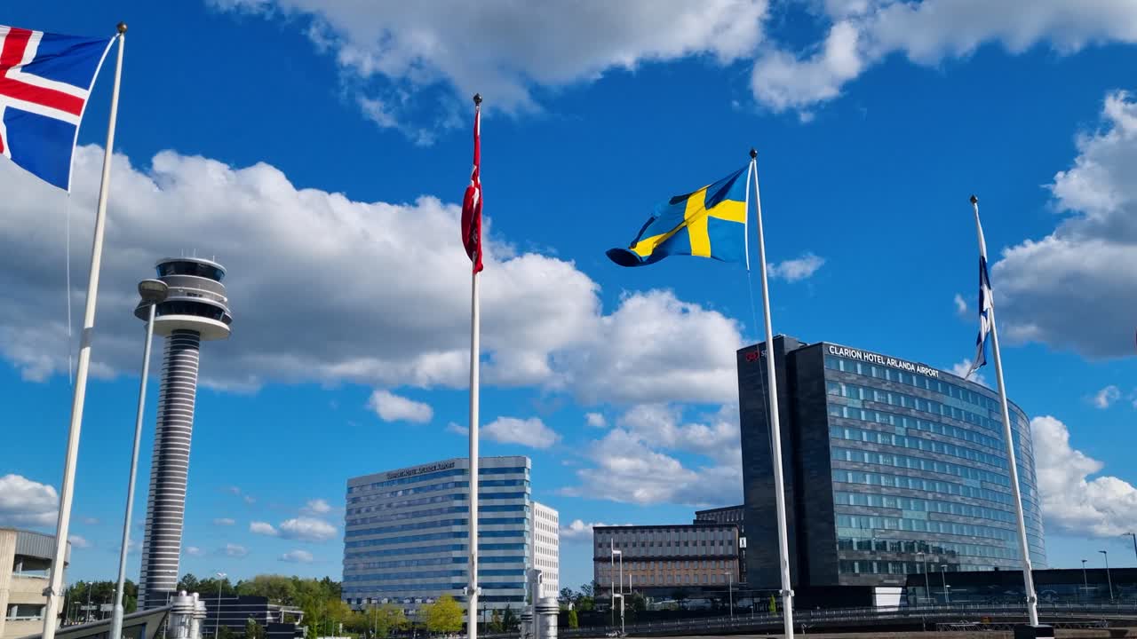 The European Union and various Scandinavian flags sway gently in the wind at Stockholm’s Arlanda Airport, with a clear blue sky framing the airport hotel and control tower