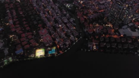 Vuelo-Nocturno-Sobre-El-Río-Y-La-Zona-Suburbana-Con-Instalaciones-Deportivas,-Tejados,-Carreteras-Con-Equilibrio-De-Luz-Ambiental-Y-Artificial