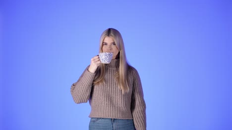 portrait of pretty blonde woman drinking from coffee mug, looking at camera against blue background, studio shot