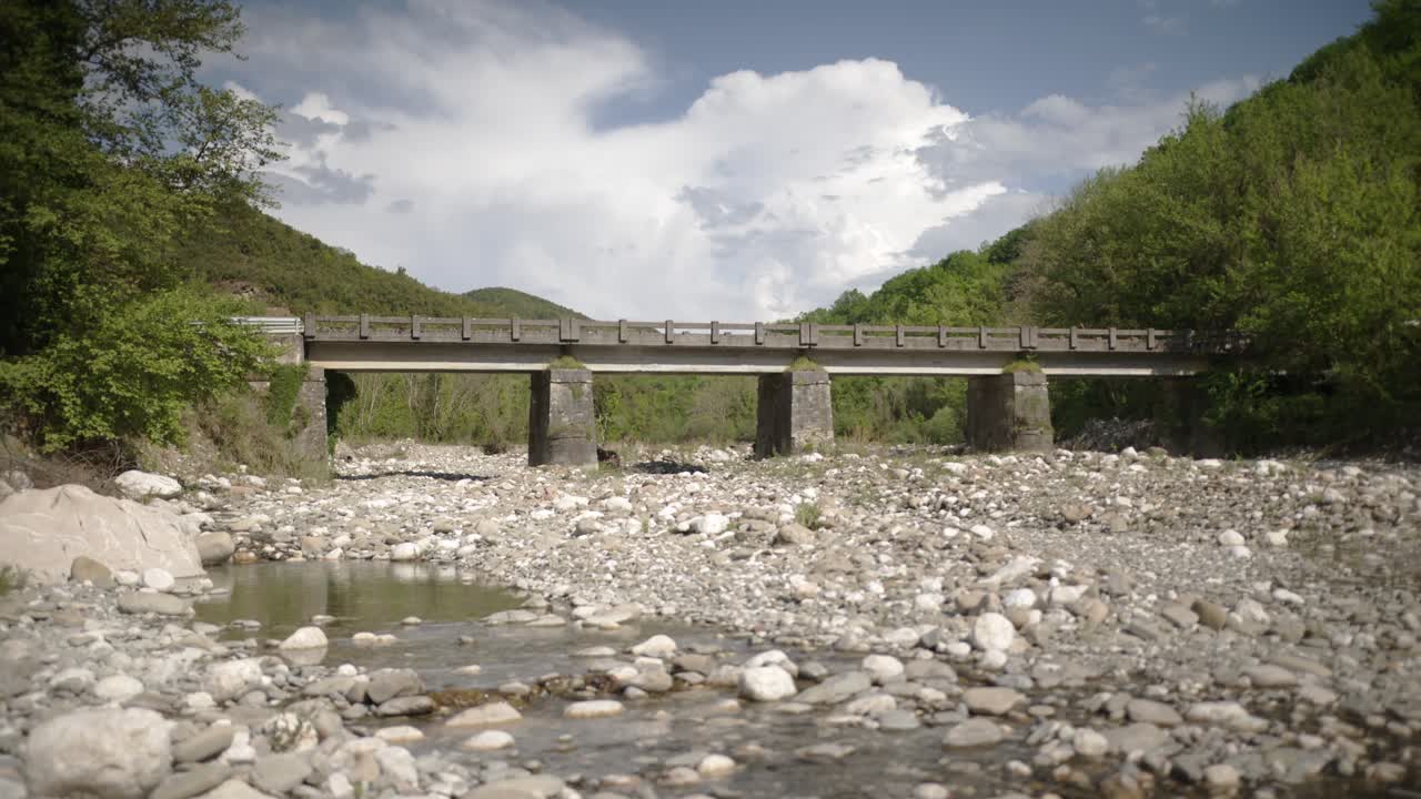 Static wide shot of a concrete bridge crossing a rocky, shallow section of the Arachthos River in Tzoumerka. Surrounded by green hills under a partly cloudy sky.