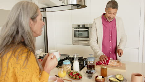 multiracial senior couple , preparing breakfast, cutting bread, drinking coffee in kitchen, at home