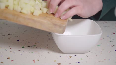 Person-moving-diced-potato-from-wooden-cutting-board-to-white-bowl,-CLOSEUP