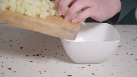 person moving diced potato from wooden cutting board to white bowl, closeup