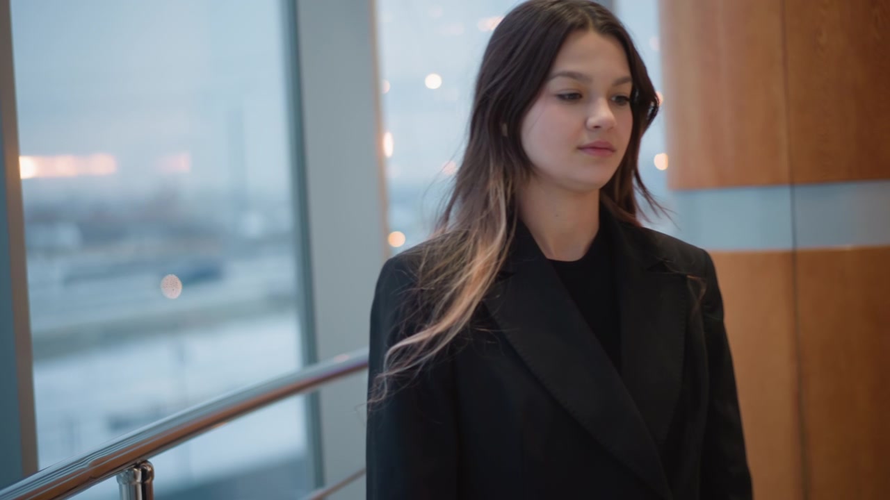 Giovane donna ride mentre cammina accanto a una grande finestra luminosa all'interno di un interno moderno, indossando un blazer con i capelli che le fluiscono e le spalle rilassate, mostrando un'espressione vivace, un movimento naturale e un'energia positiva.