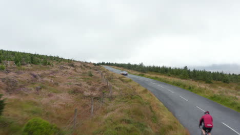 Radfahrer-Und-Autos-Auf-Der-Bergstraße,-Die-Den-Hügel-Hinaufführt.-Wald-Mit-Jungen-Bäumen-Entlang-Der-Straße.-Bedeckter-Himmel.-Irland