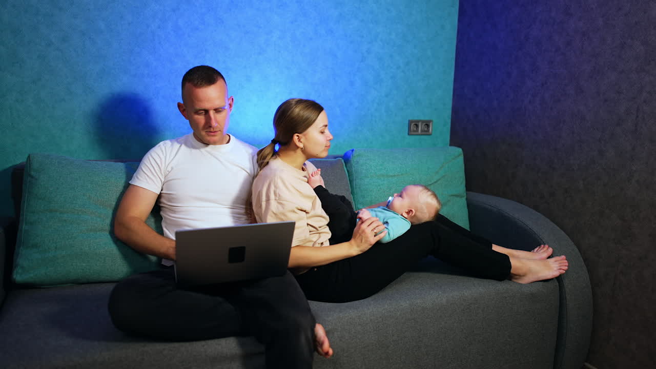 Woman sits on the sofa leaning on her husband and holding a baby on her knees. Man sits with laptop on his laps focused on screen.