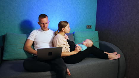 woman sits on the sofa leaning on her husband and holding a baby on her knees. man sits with laptop on his laps focused on screen.