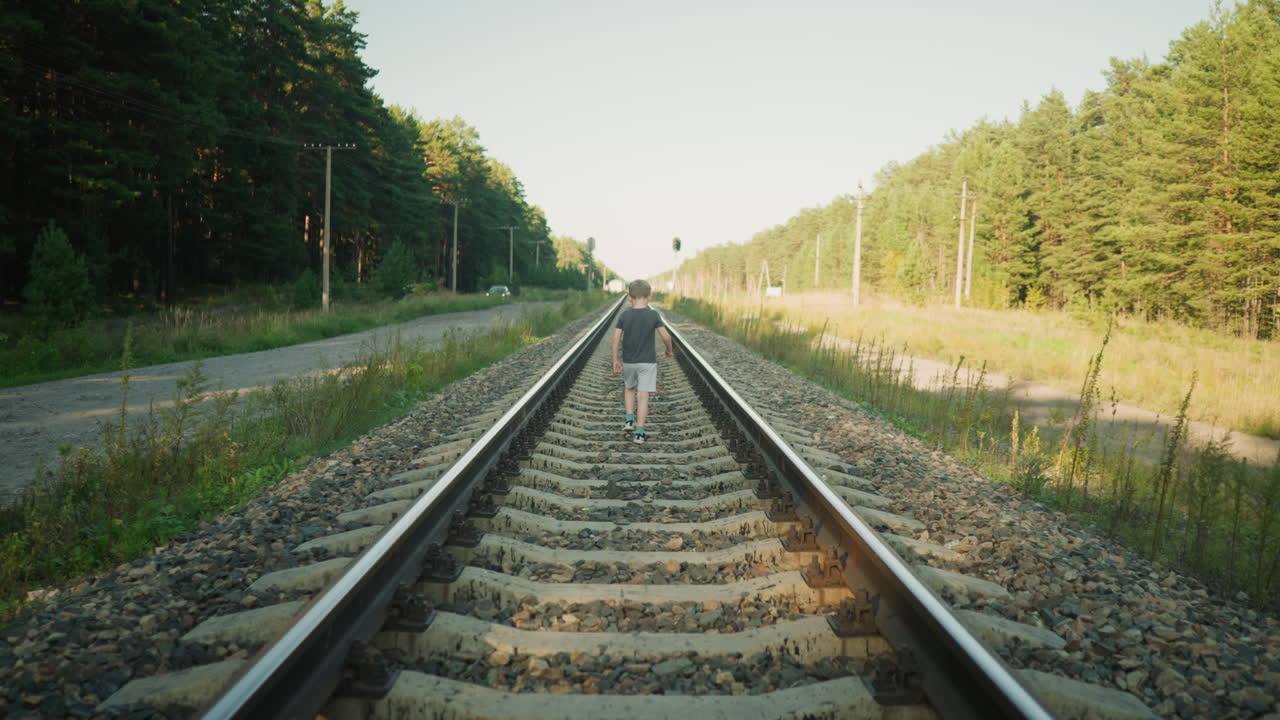back view of boy strolling along railway track balancing on gravel ties with arms outstretched under bright sunlight, surrounded by forest scenery and power line poles lining rural path