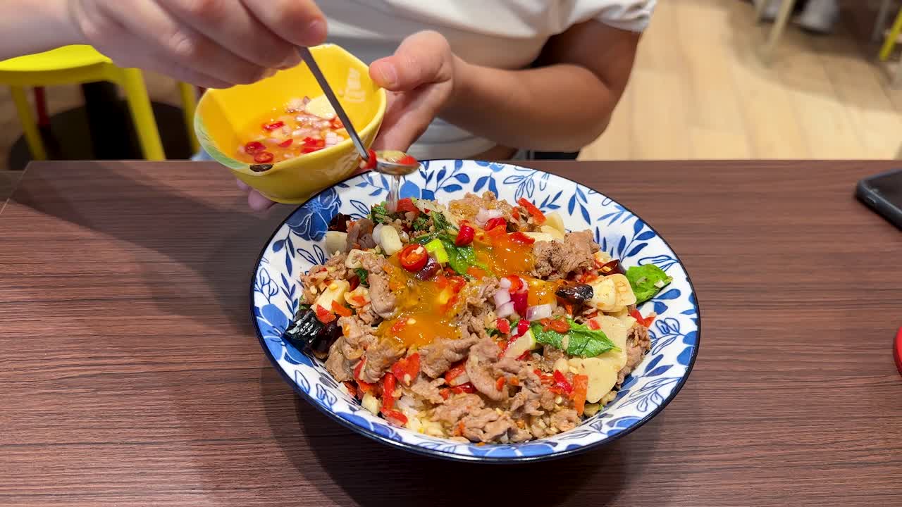 Pouring chili garlic fish sauce onto beef rice bowl in a bright, casual Asian restaurant