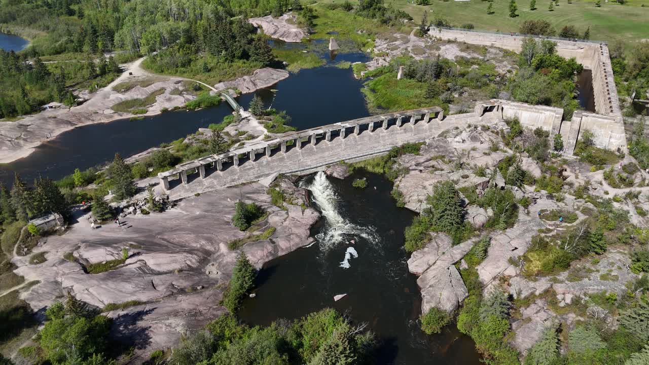 Drone che guarda in basso e poi si muove verso l'alto mentre avanza per rivelare la diga Old Pinawa a Manitoba, mostrando acqua impetuosa, la struttura in cemento e le cime degli alberi verdi.