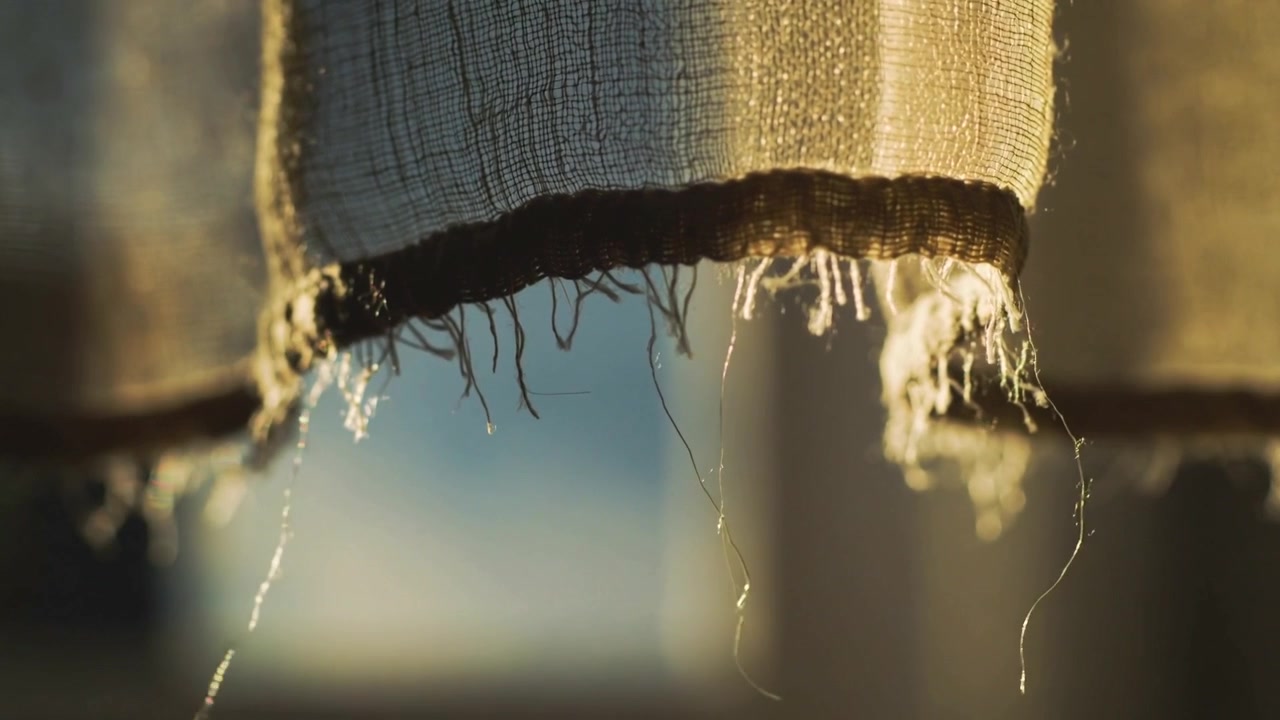 Orlo di tenda tessuta che ondeggia con fili sfilacciati alla finestra illuminata dal sole, una leggera brezza che spinge le goccioline verso il basso.