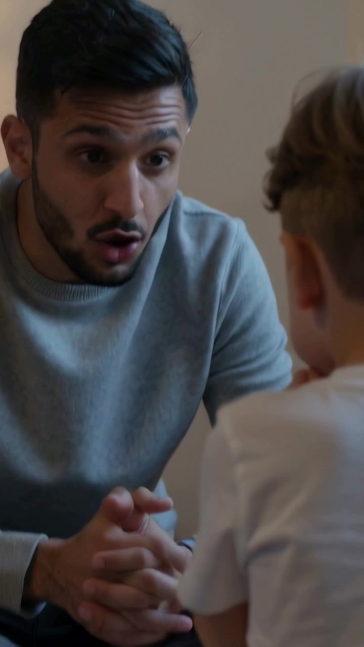 Video verticale: Padre che parla si china in avanti, cercando di coinvolgere il figlio sul tappeto, maglione grigio.