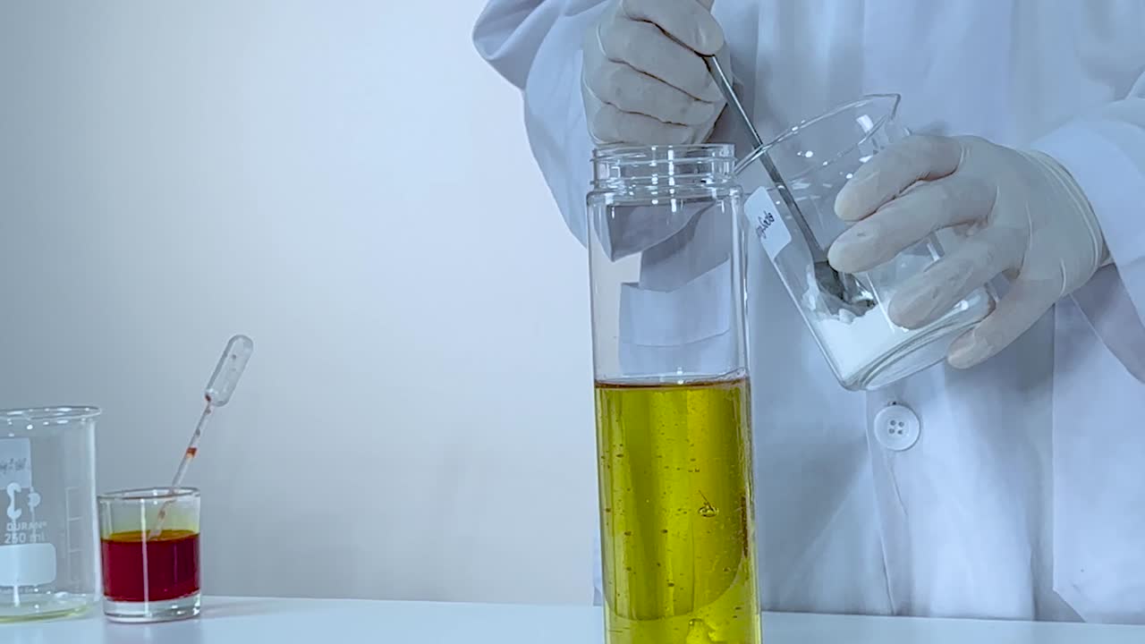 A scientist mixes white powder into a tall cylinder of yellow liquid using a spoon.