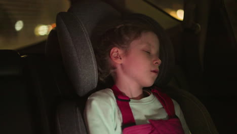 una vista de cerca de un niño durmiendo pacíficamente en un asiento de coche, iluminado por el brillo suave de las luces ambientales, el niño lleva un mono rosa y una camisa verde claro, descansando cómodamente