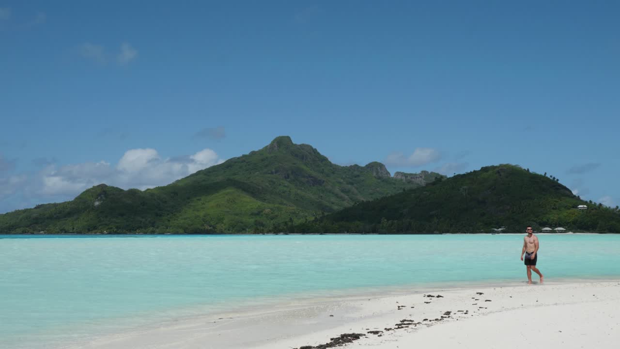 Young caucasian man in a swimming suit and sunglasses walking on a tropical pacific white sand beach on a sunny day with a small green island in background in Maupiti, French Polynesia.