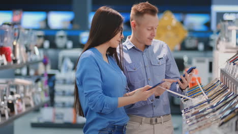 Man-holding-smart-phone-and-pointing-on-other-phone-which-is-in-woman-hands.-Tech-store-interior.