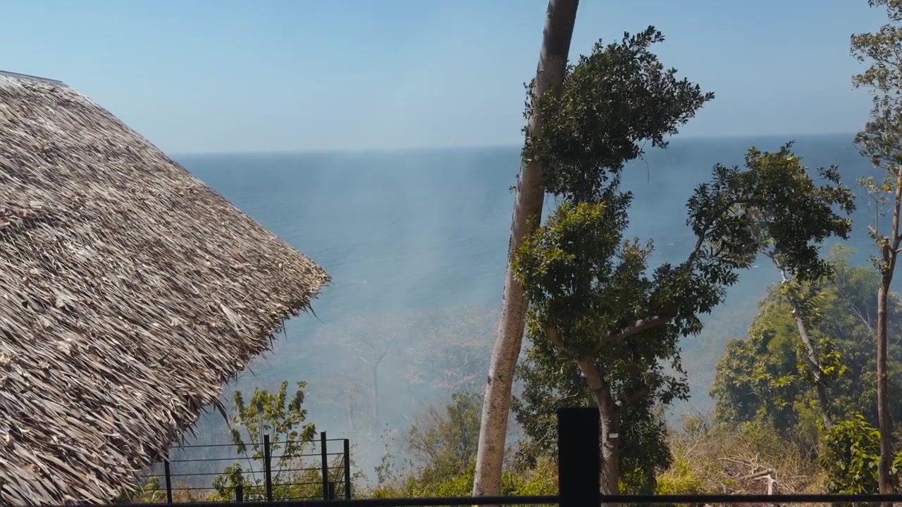 Inquadratura statica di fumo che si diffonde su acque calme e blu dell'oceano con tetto di paglia e alberi verdi in un ambiente costiero tropicale. · 4K DCI 25fps