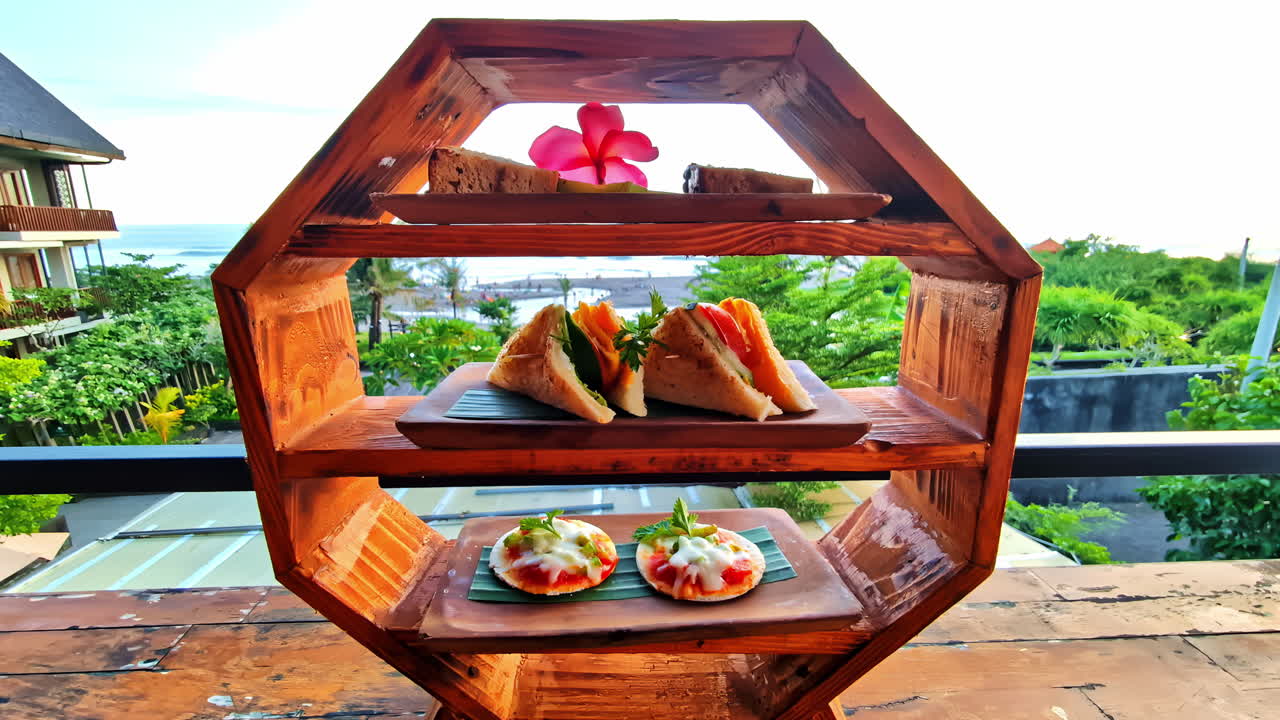 An elegant breakfast and afternoon tea, with sandwiches and pastries on a unique wooden stand, served on a luxury hotel balcony with a beautiful tropical beach view in Bali