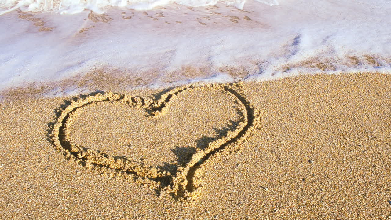 Un cuore disegnato sulla sabbia. Il cuore viene cancellato da un'onda marina. Riprese sulla spiaggia.