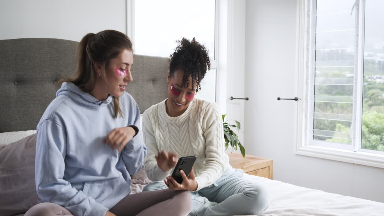 Diverse female friends scrolling smartphone tutorial on bed then peeling off pink under-eye patches