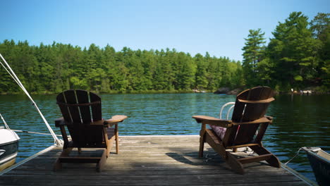 two empty muskoka chairs sit at the end of a dock looking out at the water on a calm, beautiful summer day