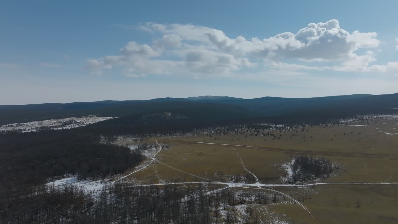 Vidstrakt luftlandskab af åben eng, nøgen skov og snoede grusveje med sneflekker under dramatiske skyer, roligt landskab om vinteren