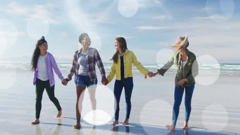 spots of light against group of diverse female friends holding hands walking on the beach