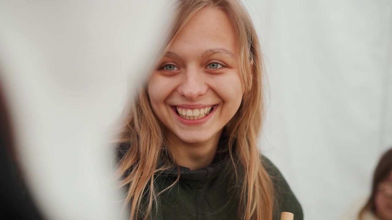 Ritratto a mezzo busto di una giovane donna bionda che passa da uno sguardo pensieroso a un sorriso gentile e a una risata aperta, mentre si protende verso i compagni accanto a una tenda da esterno, creando un momento sociale spontaneo e amichevole.