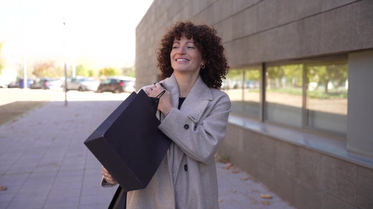 Una donna elegante con i capelli ricci, in un contesto urbano, tiene una borsa della spesa con articoli deliziosi.