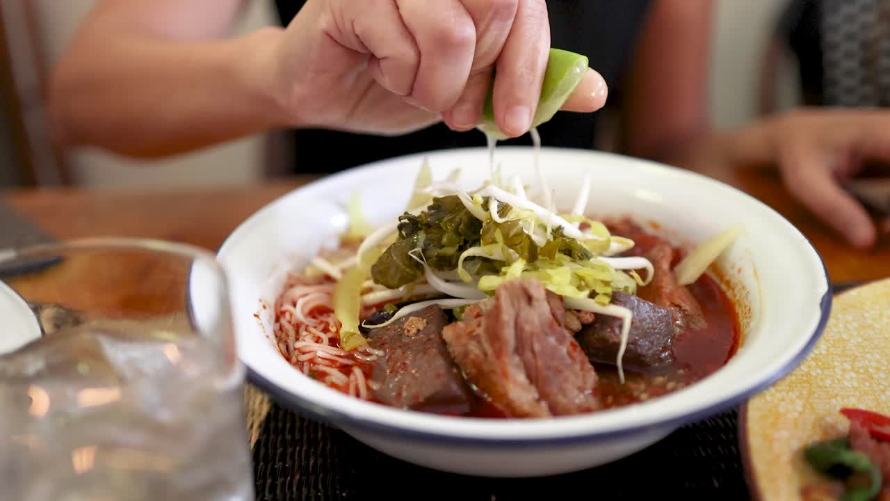 A hand squeezes fresh lime over a bowl of rice vermicelli with Northern Thai curry sauce, enhancing vibrant ingredients in a warmly lit dining setting