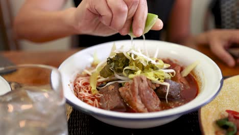 a hand squeezes fresh lime over a bowl of rice vermicelli with northern thai curry sauce, enhancing vibrant ingredients in a warmly lit dining setting