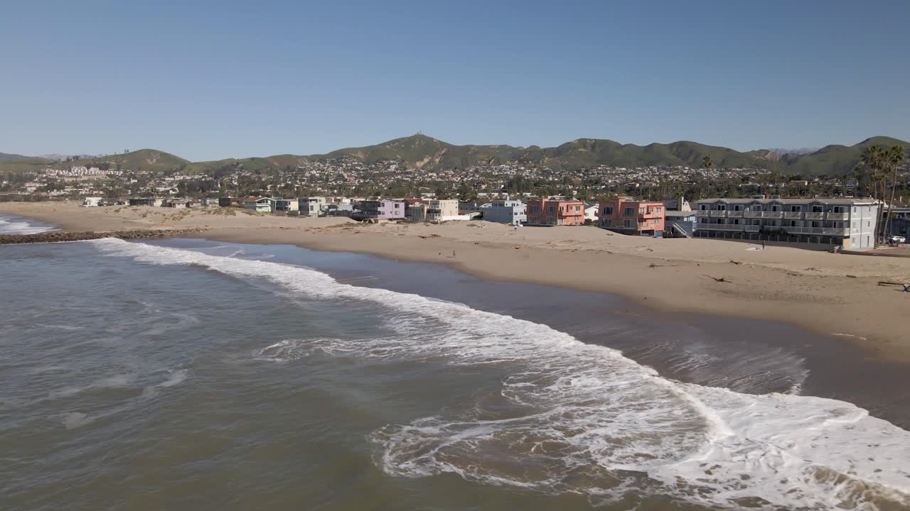 Low drone flyby capturing Ventura Beach's calm waves, sandy shore, beachfront homes, and glimpses of Ventura city