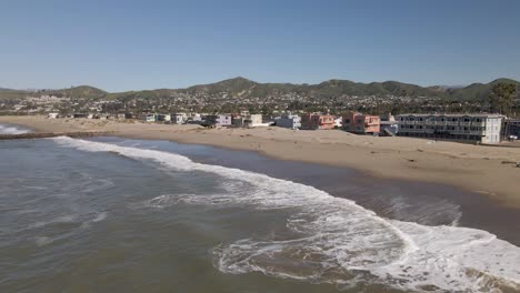 Low-drone-flyby-capturing-Ventura-Beach's-calm-waves,-sandy-shore,-beachfront-homes,-and-glimpses-of-Ventura-city