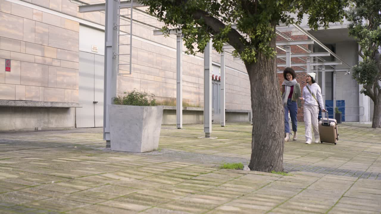 Female friends entering courtyard pulling tan and dark suitcases past tree under canopy, copy space