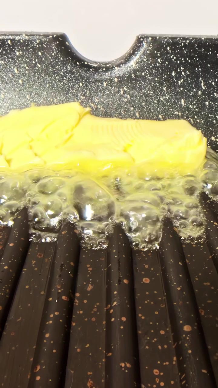 A stick of butter melts and sizzles on a hot griddle, creating bubbles.
