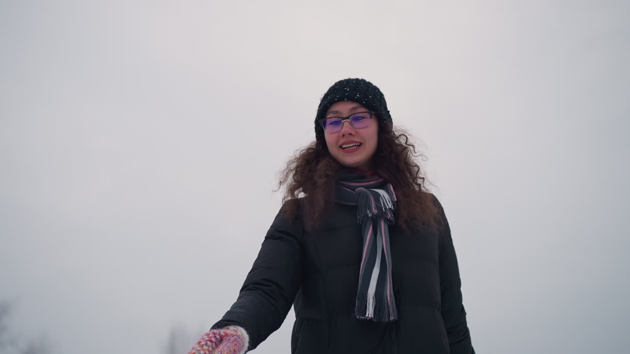Lady skating outdoors in winter snow wearing black coat, knit hat, striped scarf and colorful gloves, enjoying seasonal activity with graceful expression under cloudy sky creating peaceful atmosphere